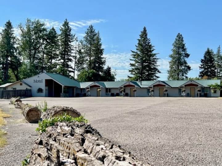 Motel row in Glacier National Park with a gravel lot, low buildings and tall evergreen trees under a bright blue sky.