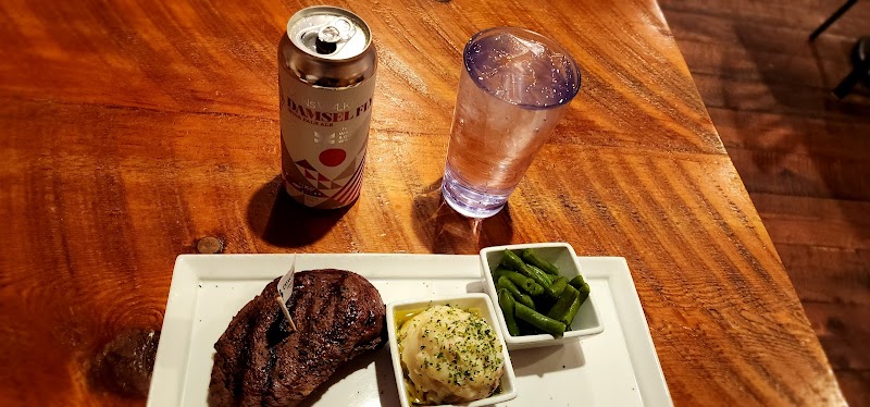 Wooden table in Yellowstone National Park shows a grilled steak, mashed potatoes, green beans, beer can and water glass.
