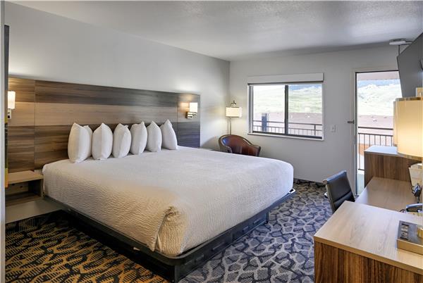 Spacious Yellowstone National Park hotel room with a king bed, white quilt, multiple pillows, desk, chair, and balcony view.