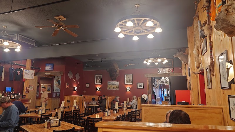 Rustic indoor dining room in Yellowstone National Park with wooden tables, patrons eating, mounted animal heads, and wagon-wheel chandeliers.