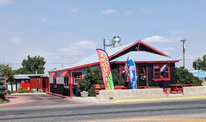 Colorful roadside restaurant with red and black siding, gray metal roof, flags, and a small patio in Glacier National Park.