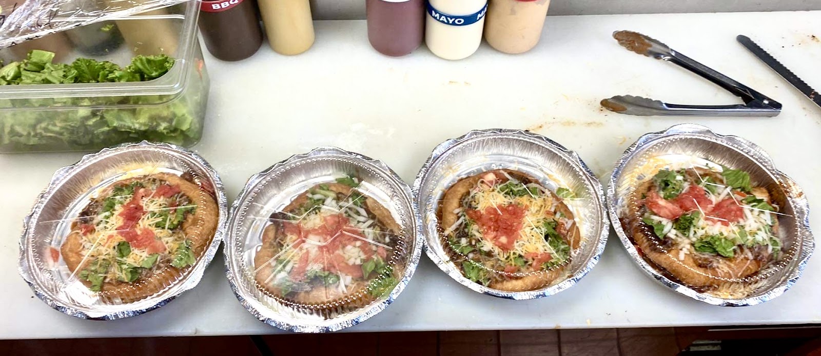 Four foil-wrapped personal pizzas topped with lettuce, tomato, onion, cheese, cilantro on a prep table in Glacier National Park.