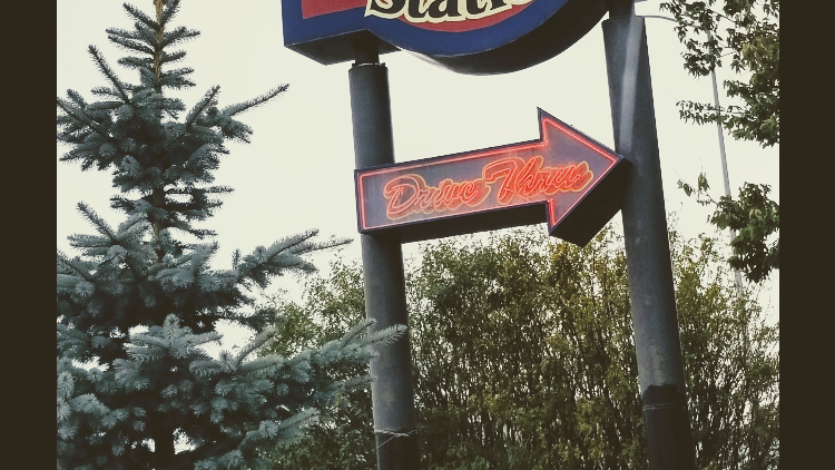 Neon Drive-Thru arrow sign on tall dark posts beside pine trees in Glacier National Park.