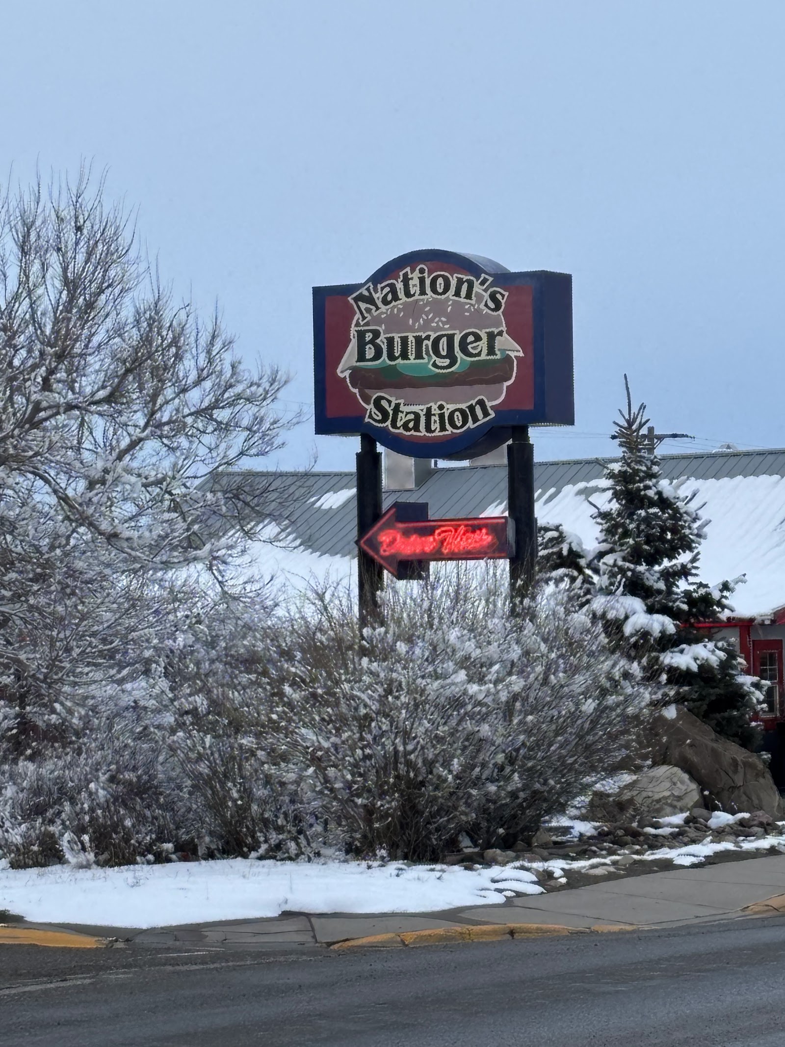 Snowy roadside in Glacier National Park with a large burger sign and red neon arrow beside snow-dusted shrubs and a building.