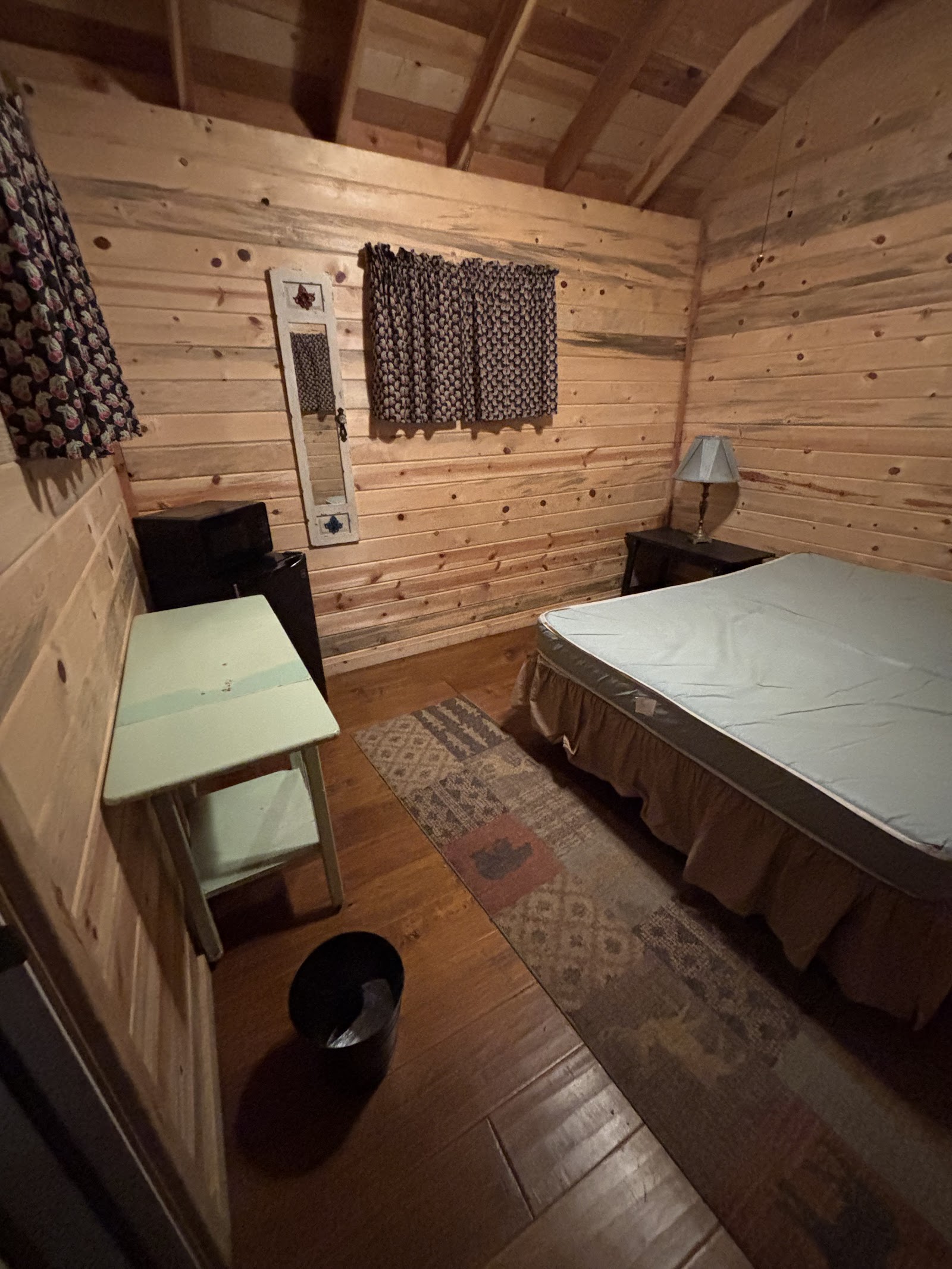 Cozy log cabin bedroom in Glacier National Park with a full bed, patterned curtains, a small green desk, mirror and lamp.