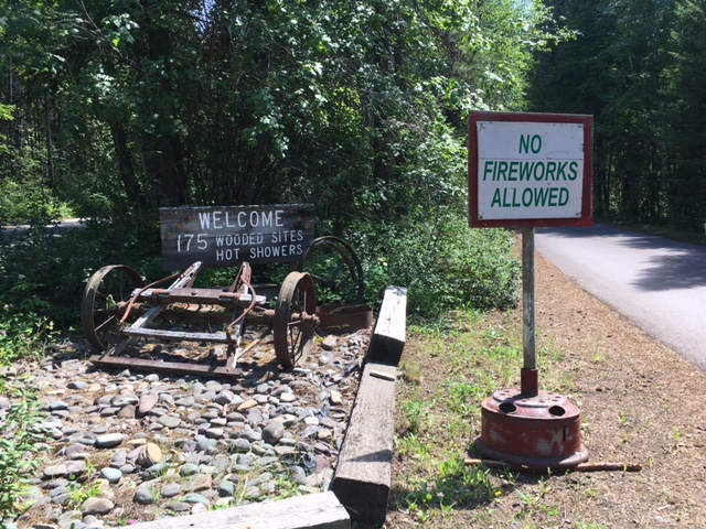 Glacier National Park: rusted wagon wheel display beside a rocky bed and a No Fireworks Allowed sign by a forest road.