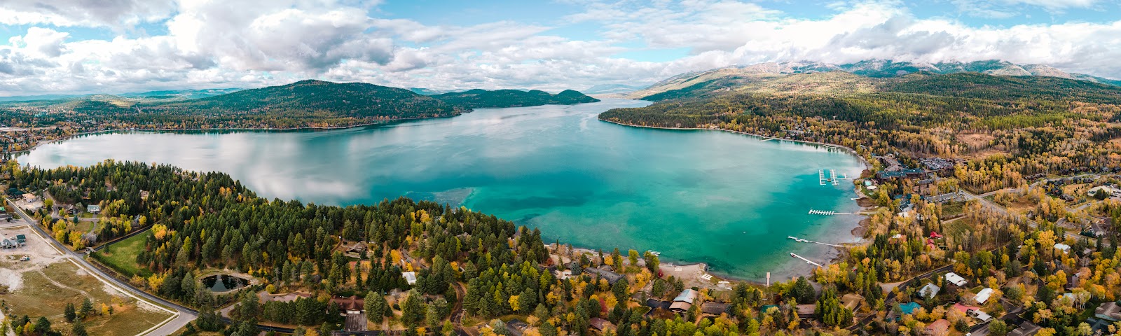 Aerial view of a turquoise glacier lake bordered by pine forests and a lakeside town with docks in Glacier National Park.