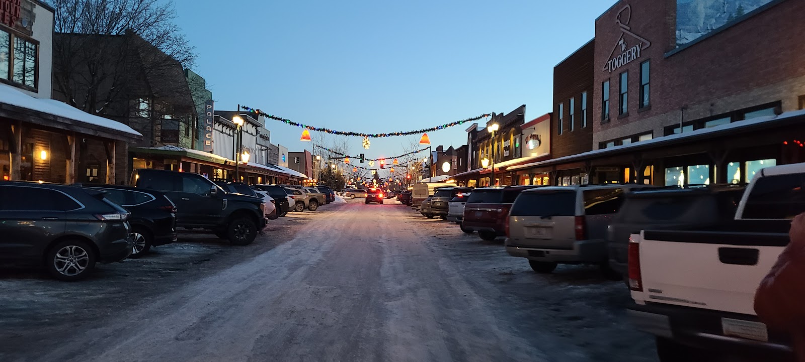Snowy Main Street in Whitefish, Glacier National Park, lined with parked cars, string lights, and storefronts at dusk.