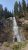 Waterfall cascading down a rocky cliff at Whitefish in Glacier National Park, framed by tall evergreen trees.