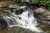 Hidden Falls cascades over dark rocks with green moss and foam-filled pool in Glacier National Park.