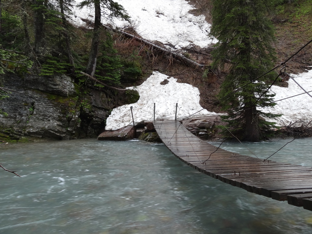 Suspension wooden bridge curves over a turquoise river in Glacier National Park, with snow-dusted banks and evergreen trees.