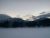 Snowy Lake Five in Glacier National Park, a frozen lake with dark pines and distant snow-dusted mountains at dusk.