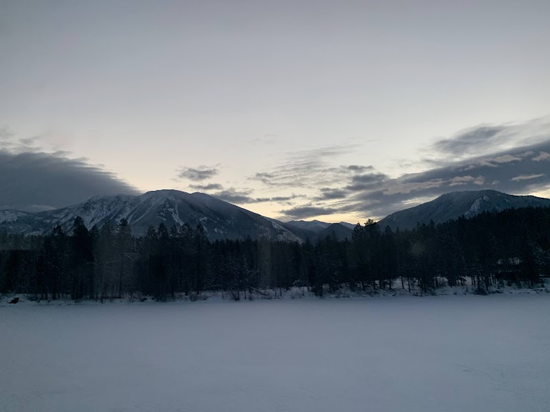 Snowy Lake Five in Glacier National Park, a frozen lake with dark pines and distant snow-dusted mountains at dusk.