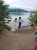 Couple by Lake Five in Glacier National Park pose on a rocky shore; bride in a white gown, groom with hat and suspenders holding a bottle while a friend waves.