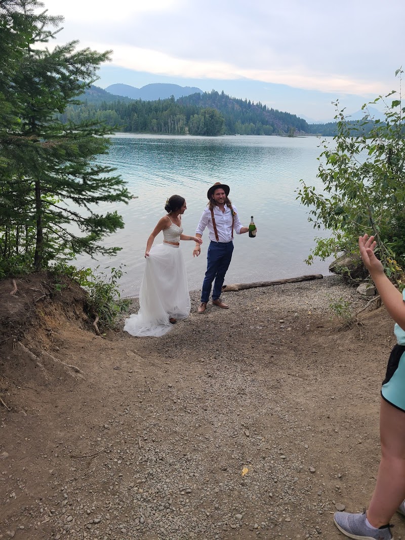 Couple by Lake Five in Glacier National Park pose on a rocky shore; bride in a white gown, groom with hat and suspenders holding a bottle while a friend waves.