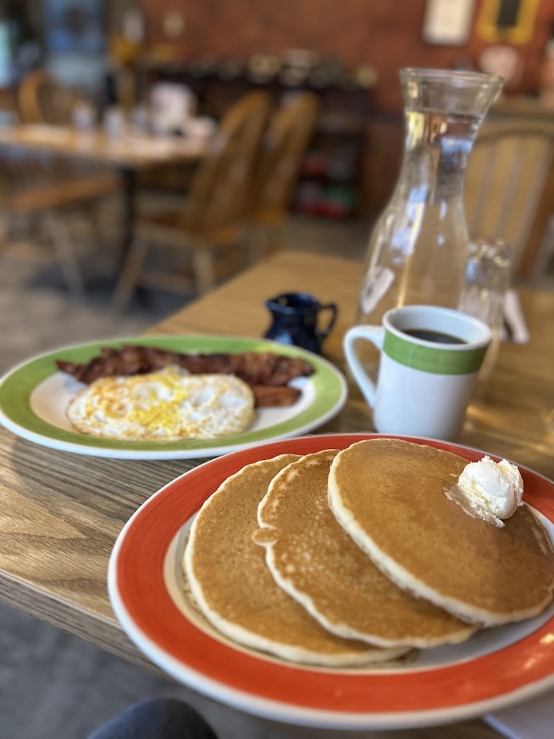 Stack of fluffy pancakes with butter on a red-rimmed plate, beside bacon and eggs, plus coffee mug and water carafe in Yellowstone National Park.