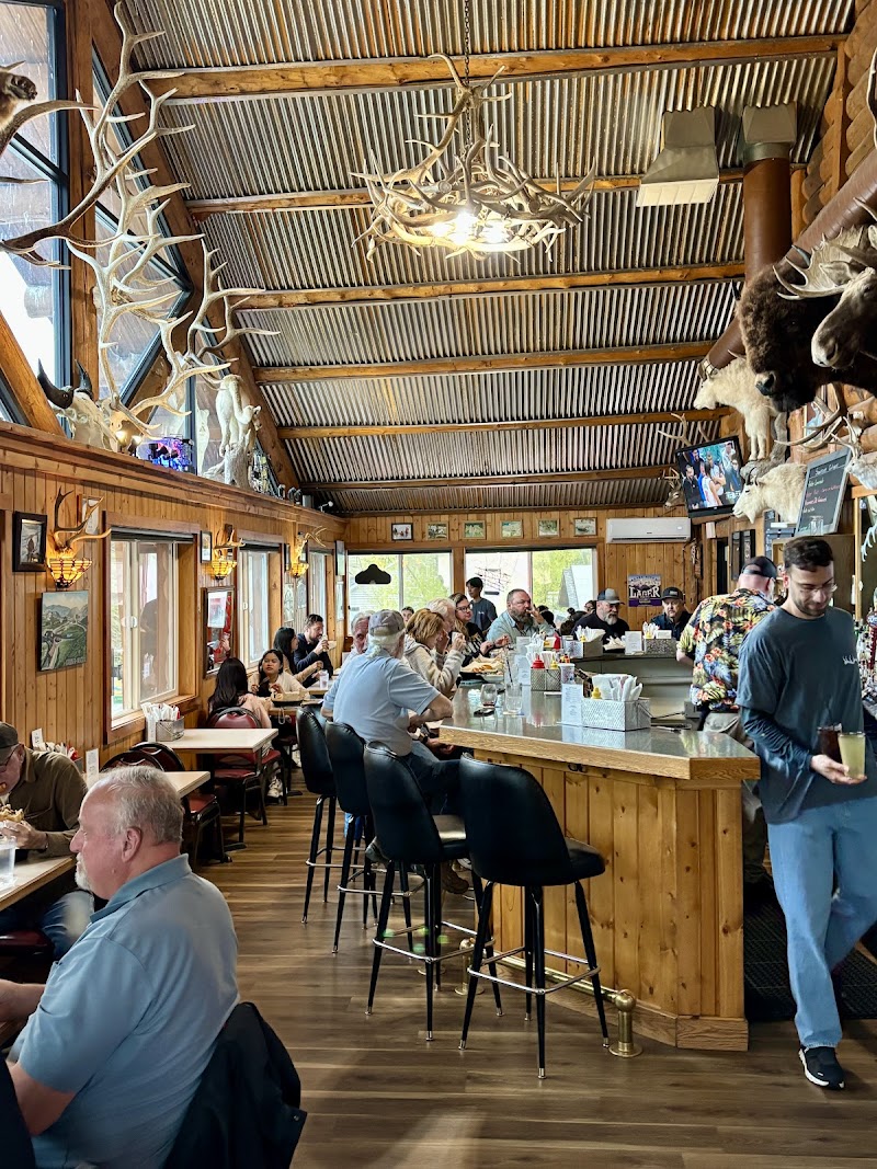 Rustic Yellowstone National Park dining room with antler chandeliers, wood walls, a long bar, and diners at tables.