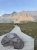 Wooden boardwalk winds through a grassy alpine meadow toward rugged glacier peaks, with a large foreground boulder and hikers nearby in Glacier National Park.