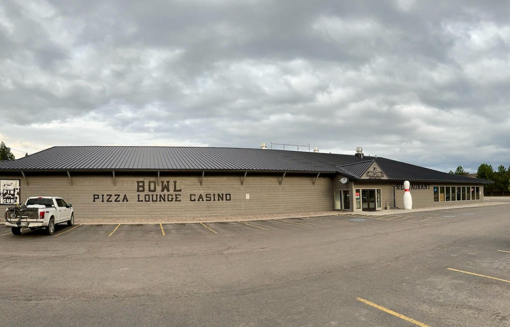 Glacier National Park exterior of Pin & Cue restaurant and bowling lounge, a low-slung building with a metal roof under a cloudy sky.