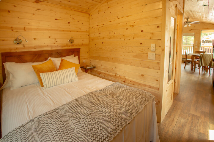 Cozy wooden bedroom in a lodge with a queen bed, yellow pillows, and a view into a dining area in Glacier National Park.