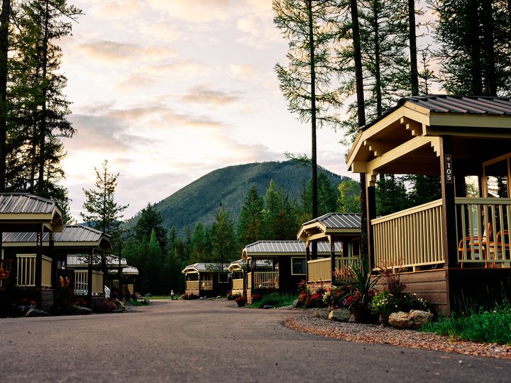 Row of wooden cabins with porches along a paved road, tall pines and a mountain backdrop in Glacier National Park.