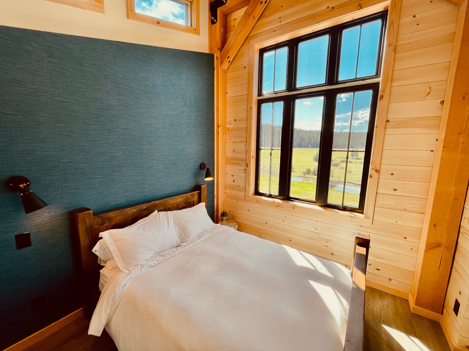 Cozy lodge bedroom with a dark blue accent wall, wooden beams, and a large window framing a valley view in Glacier National Park.