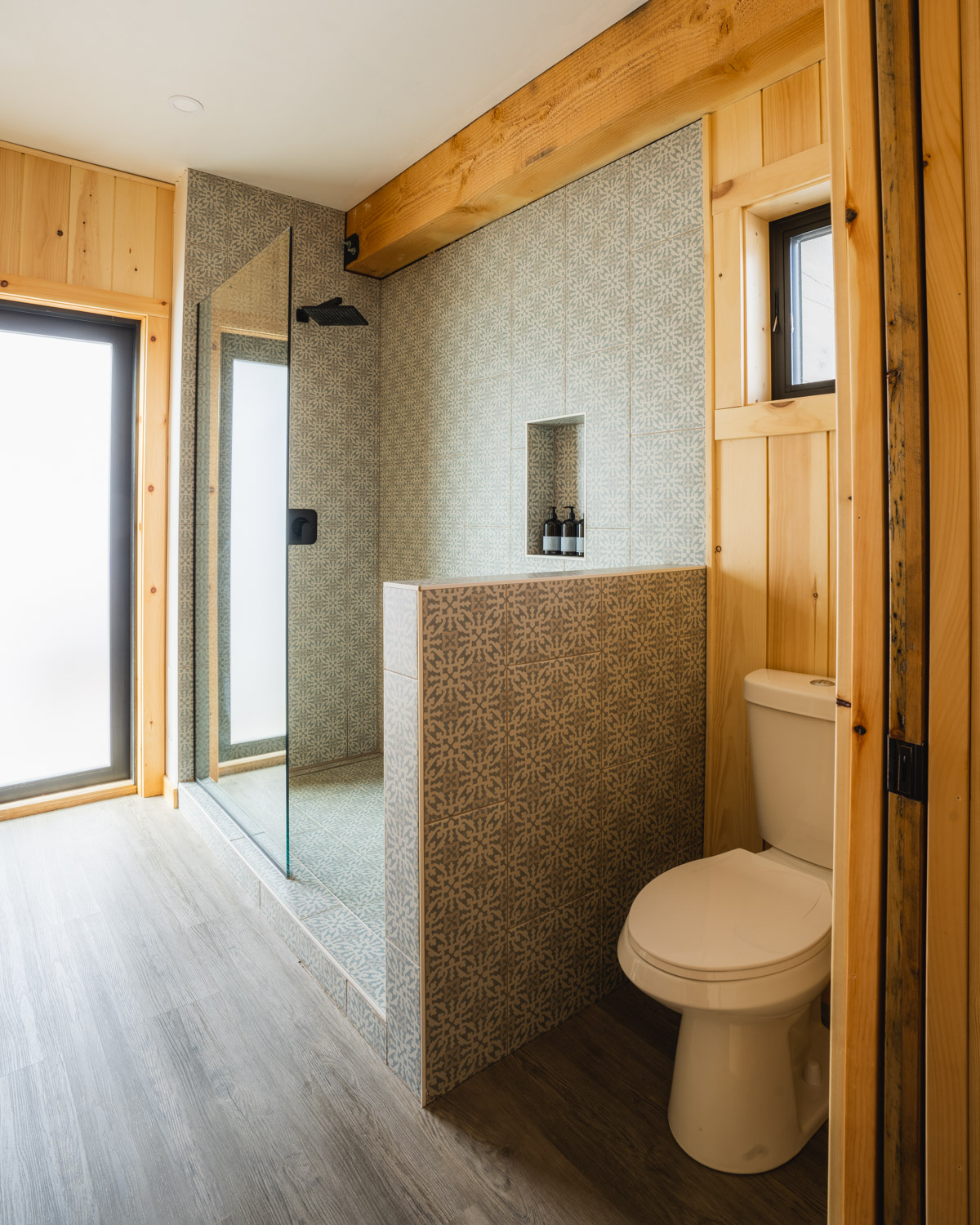 Bathroom interior at Glacier National Park lodging with wood paneling and blue patterned tile, featuring a glass shower and nearby toilet.