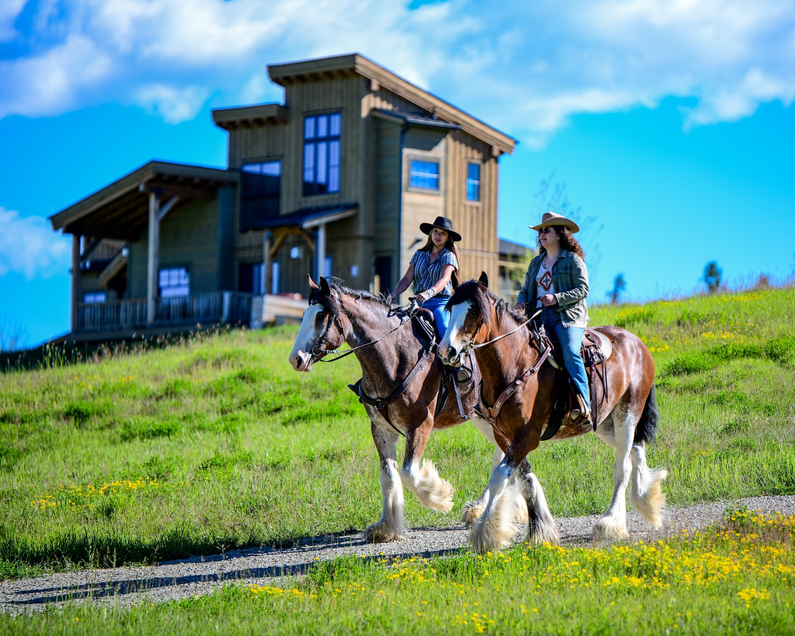 Clydesdale Outpost Guest Ranch in Glacier National Park, two riders on horseback beside a rustic lodge on a sunny hillside.