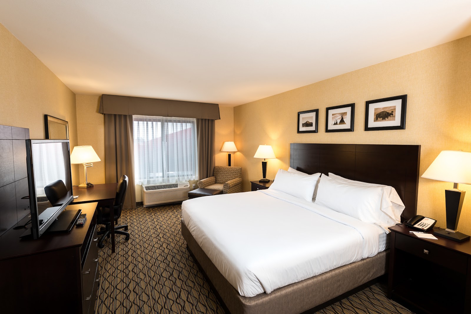 Spacious hotel room in Glacier National Park with a large white bed, dark wood headboard, framed art, nightstands, lamps, and a desk by the window.