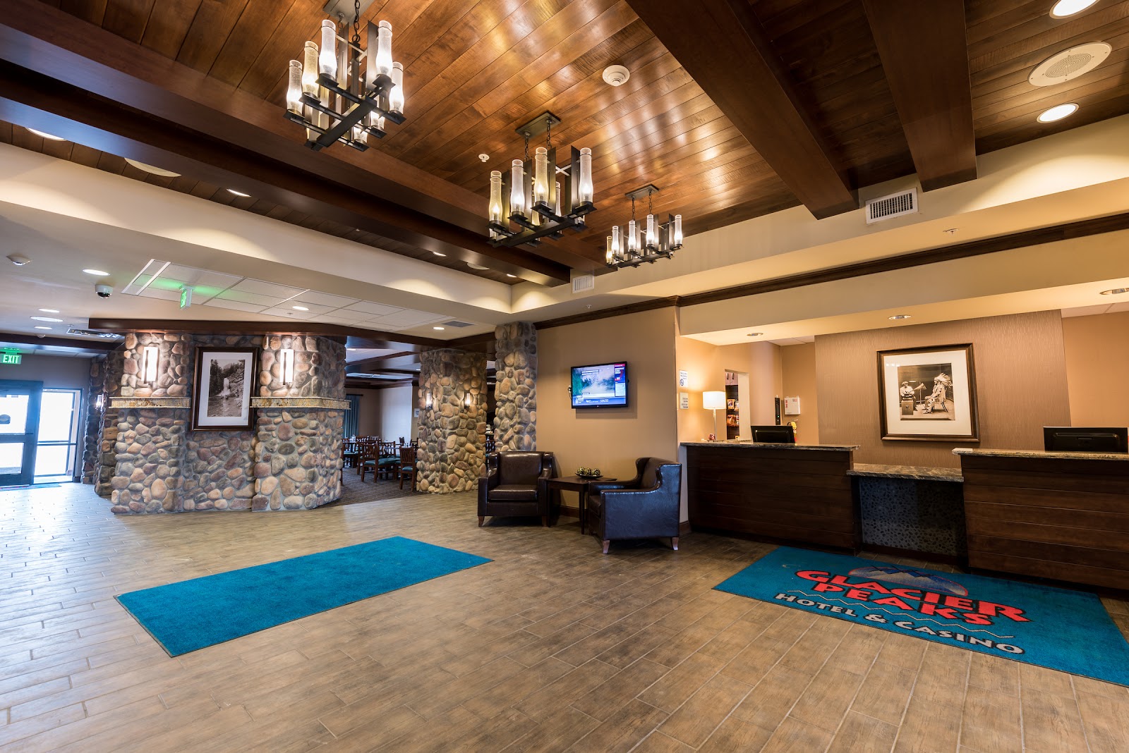 Lobby inside Glacier National Park lodging with stone pillars, dark wood beams, chandeliers, a front desk, and blue mats.
