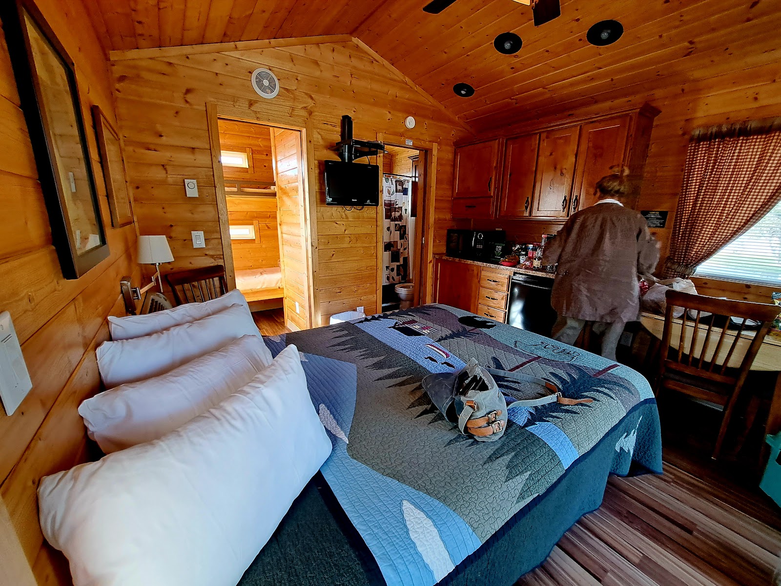 Cozy log cabin room in Glacier National Park with a quilted bed, white pillows, and a person at the kitchenette.