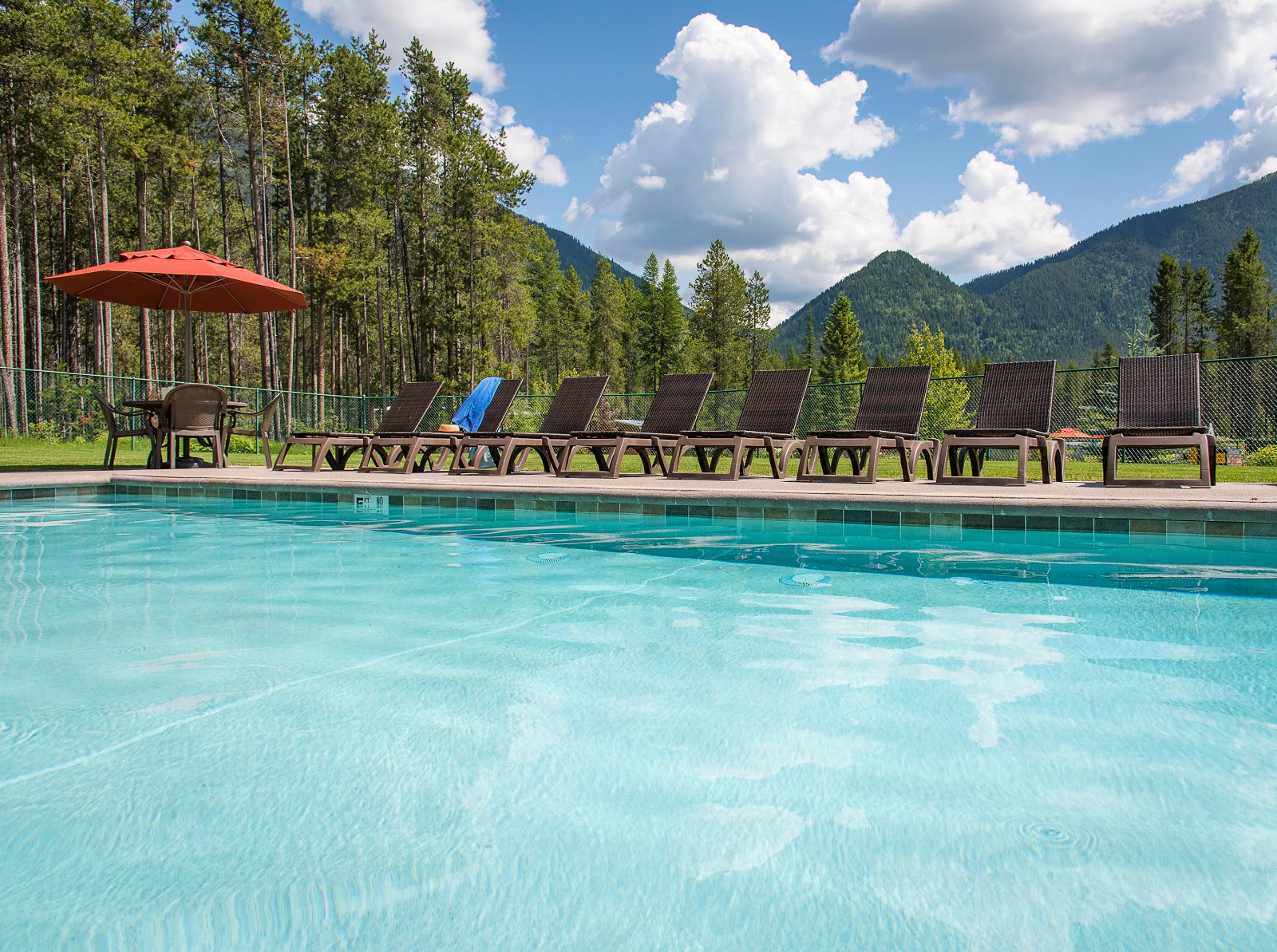 Turquoise pool with a row of brown lounge chairs and umbrella, set against pines and mountains in Glacier National Park.