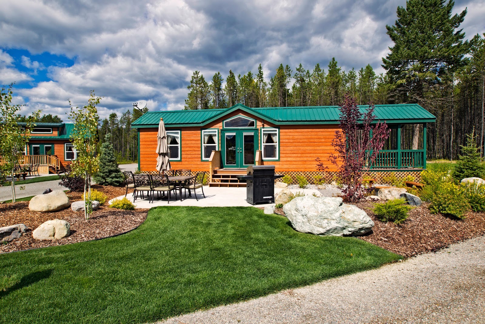 Orange cabin with green trim and a covered porch, outdoor table with umbrella, and grill in Glacier National Park campground.
