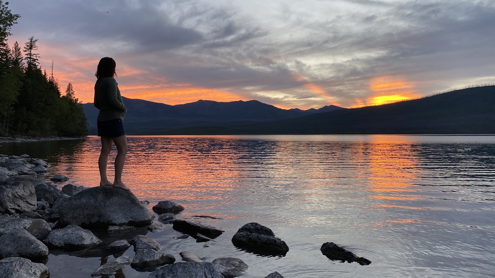 Person stands barefoot on a rocky shoreline by Glacier National Park, watching a glowing sunset reflect off the calm lake with distant mountains.