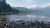 Kintla Lake shoreline with forested hills and distant Glacier peaks under a blue sky in Glacier National Park.