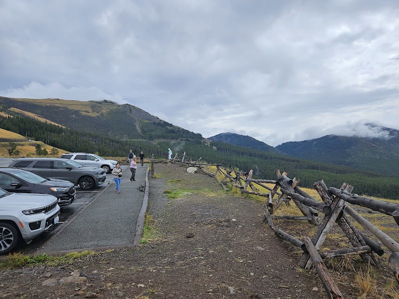 Parking lot with several cars, a gravel path and weathered wooden fence overlooking rolling forested hills in Yellowstone National Park.