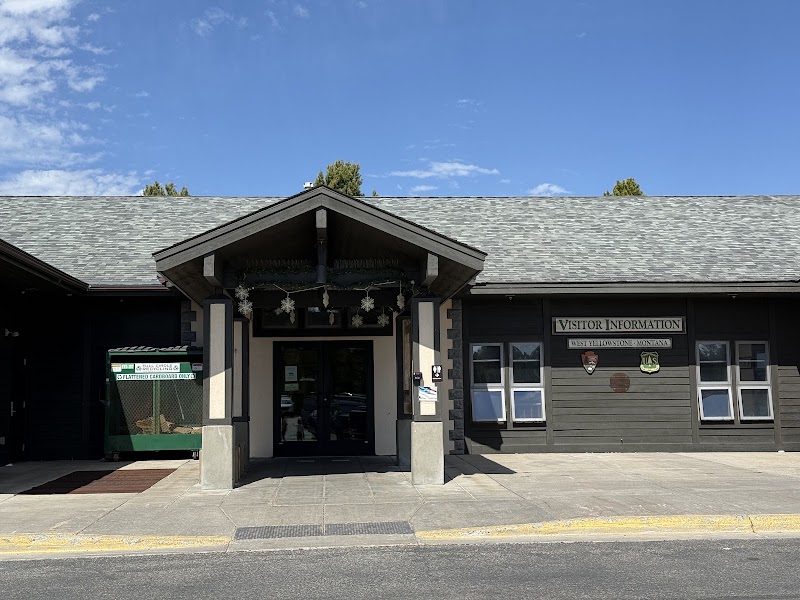 Front entrance of a visitor center with a peaked roof, gray siding, and windows in Yellowstone National Park.