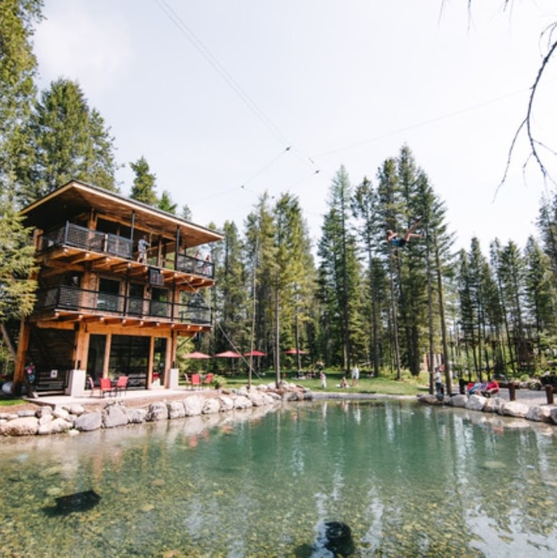 Glacier National Park scene featuring a multi-level wooden lodge beside a clear alpine pond and a zipline platform.