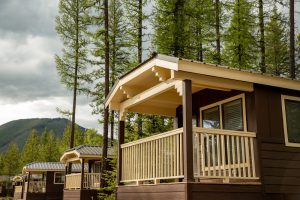 Row of brown cabins with beige porches and railings nestled among tall pines, Glacier National Park in the background.