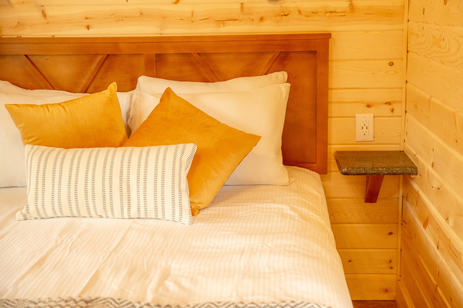 Cozy cabin bedroom with white sheets, mustard pillows and a wooden headboard, plus a granite side table in Glacier National Park.