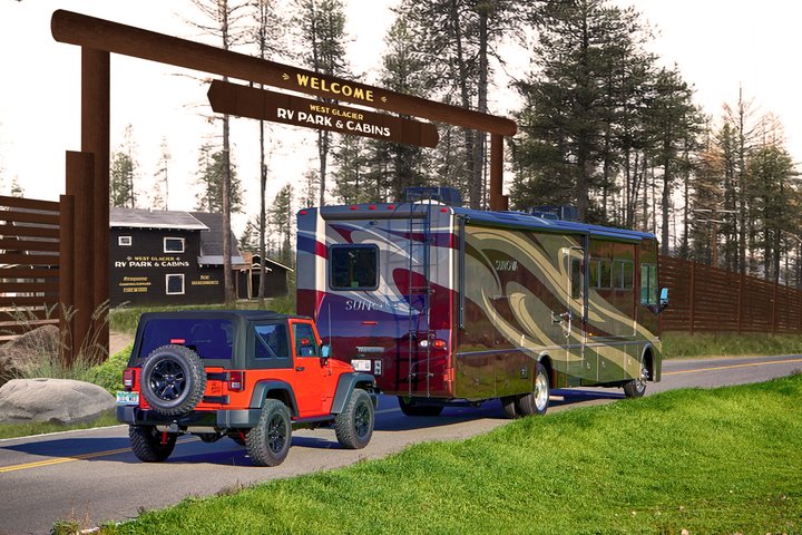 Orange Jeep with a spare tire attached to a large maroon-green motorhome beside a wooden welcome arch at Glacier National Park campground.