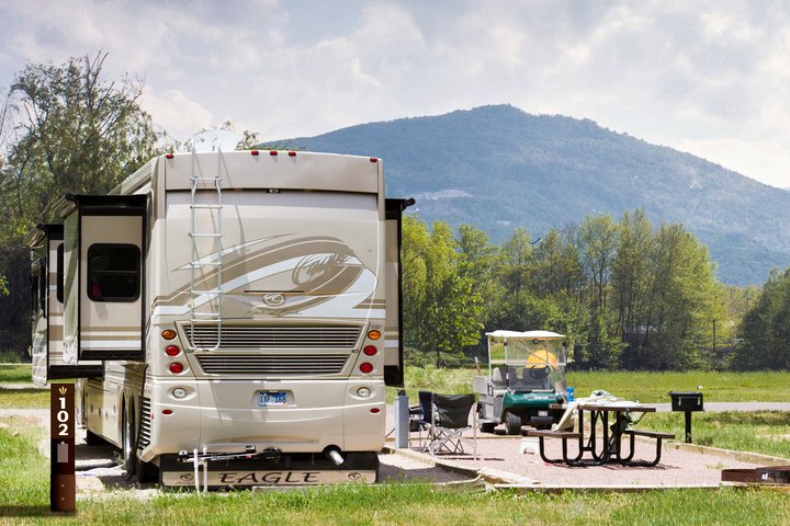 A large motorhome at a campground with a ladder on its back, a picnic table and golf cart nearby, with green fields and mountain backdrop in Glacier National Park.