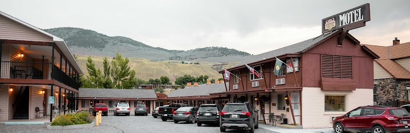 Motel-style row of buildings with a gravel parking lot, several cars, flag banners, and hills in Yellowstone National Park.