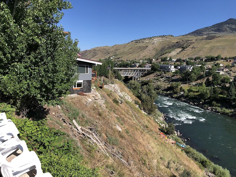 Cliffside lodging with a gray house overlooking Yellowstone National Park's river, white chairs along a path, a bridge, and rugged hills.