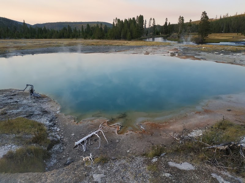 Morning Glory Pool in Yellowstone National Park mirrors a pale sky above a blue pool, with steam vents and rocky, barren rims.