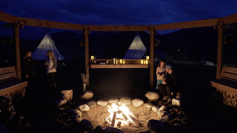 Nighttime campfire under a wooden shelter in Yellowstone National Park, with people near a row of white tipis and glowing tents.