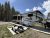 RV trailer at a campground with a wooden picnic table, blue camping chairs, a man in a red cap, and tall conifer trees in Yellowstone National Park.