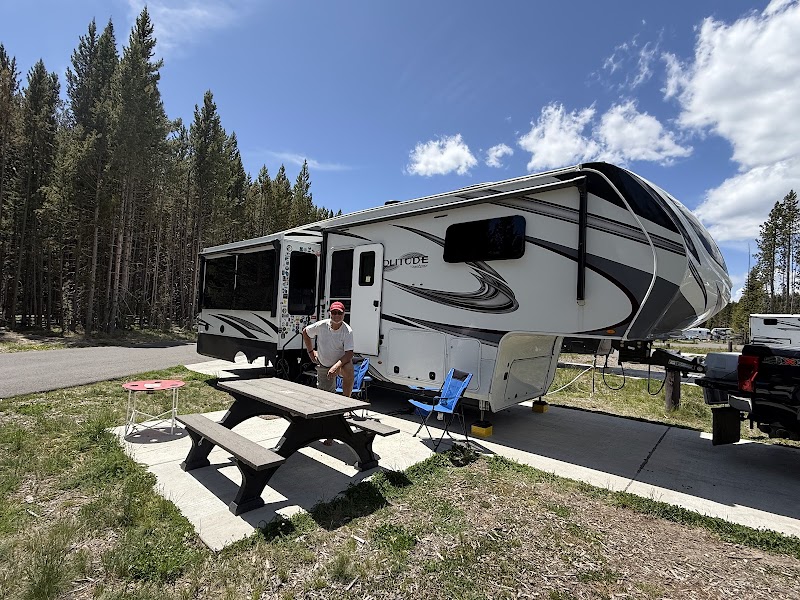 RV trailer at a campground with a wooden picnic table, blue camping chairs, a man in a red cap, and tall conifer trees in Yellowstone National Park.