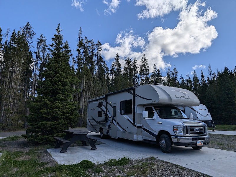 Yellowstone National Park campground shows a gray and white RV on a concrete pad beside a picnic table under tall pines.