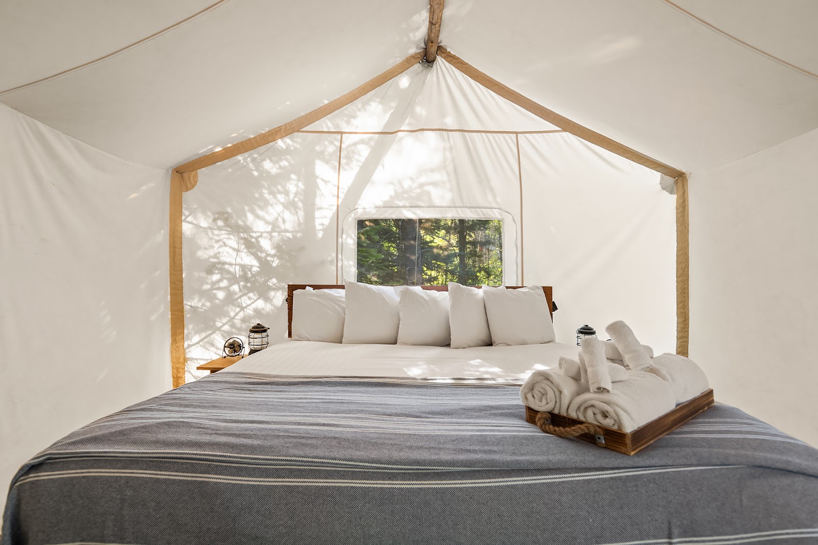 Inside a canvas tent at Glacier National Park, a neatly made bed with white pillows, gray striped blanket, lanterns, and a towel tray.